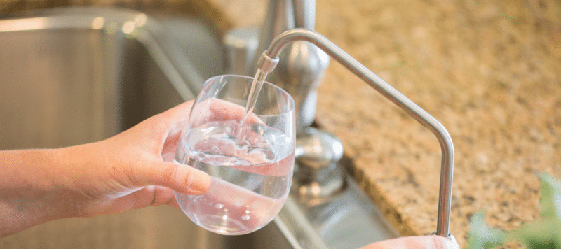 hands of a woman turning on a kitchen water faucet to fill a glass