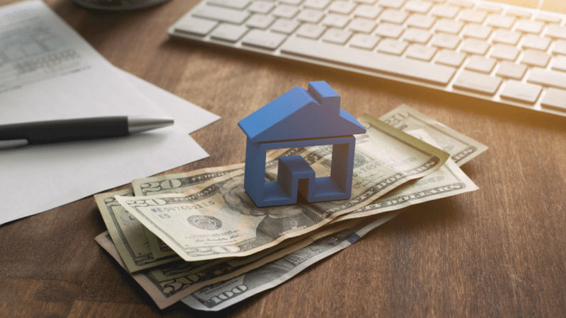 A small blue home figurine sitting on top of a stack of cash on a desk next to a keyboard and contract and pen.